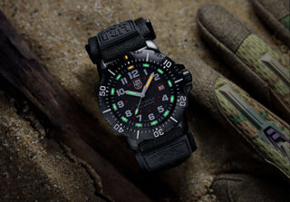 A rugged black Navy SEAL watch with a luminous dial rests on sand beside a pair of camouflaged gloves, evoking an adventurous and tactical tone.